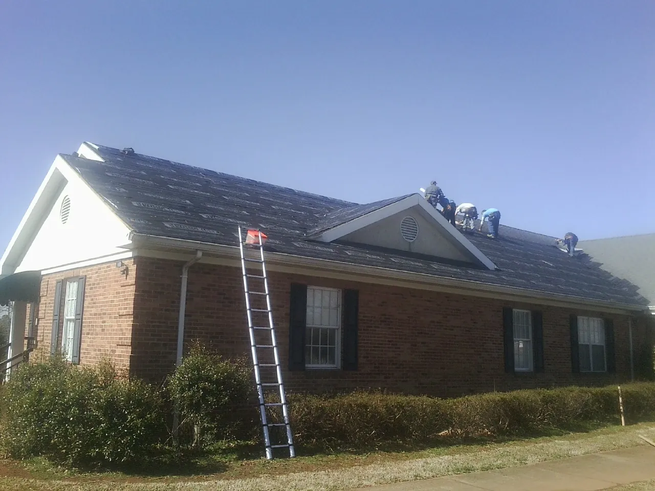 Crew tearing shingles off a brick ranch roof with a ladder set — Emergency Roof Repair work by our crew in Corfu