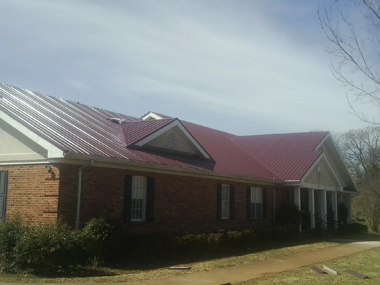 Red metal roof with gabled entries on a long brick building, from a Hail Damage Roof Repair job in Corfu