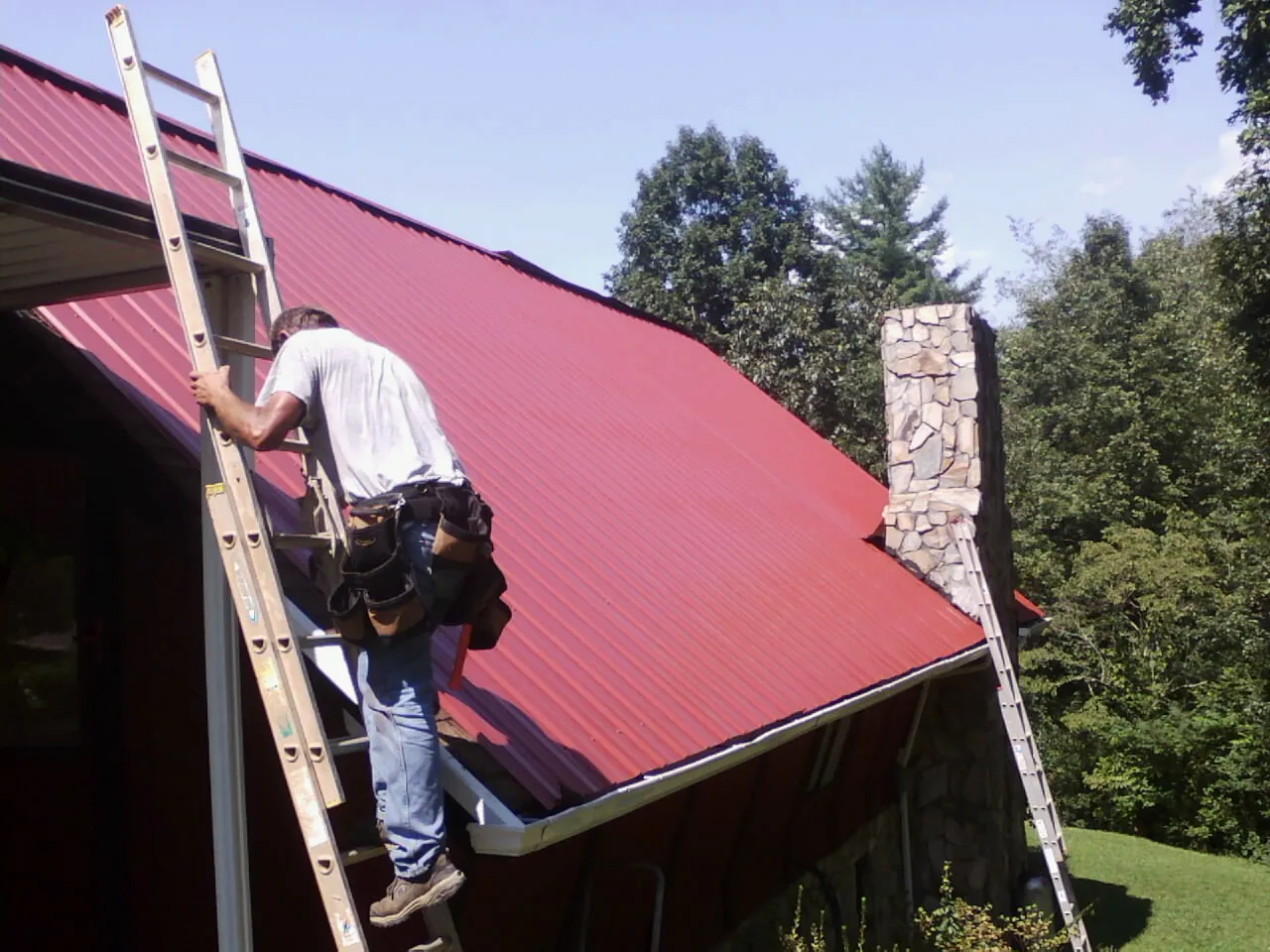 Roofer on a ladder beside a red metal roof and stone chimney — Soffit & Fascia Repair by Roofing Brothers in Corfu